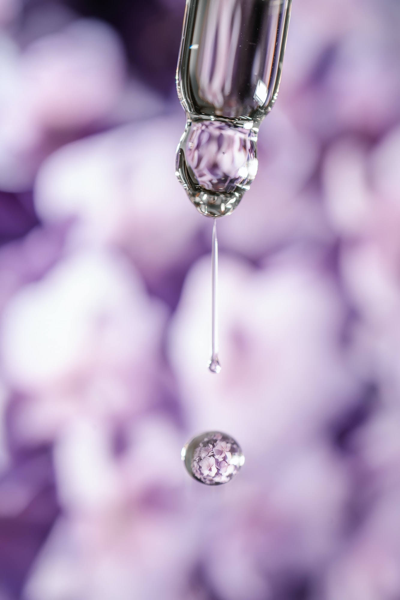 A drop of cosmetic oil falls from the pipette on a purple floral background.